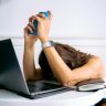 A woman overwhelmed by work, resting head on table with laptop, phone, and smartwatch.