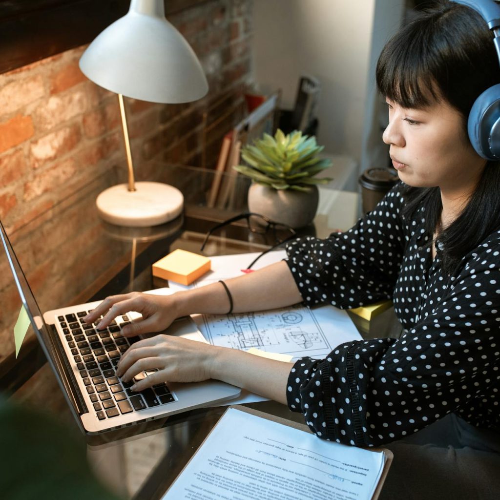 Focused woman wearing headphones, working on a laptop in a cozy home office setup with natural light.