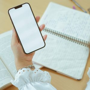 Close-up of a woman's hand holding a blank smartphone over open notebooks, ideal for mockup designs.