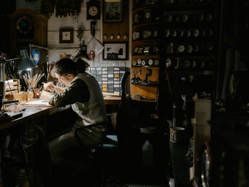 A watchmaker meticulously repairing watches in a dimly lit workshop filled with clocks and tools.